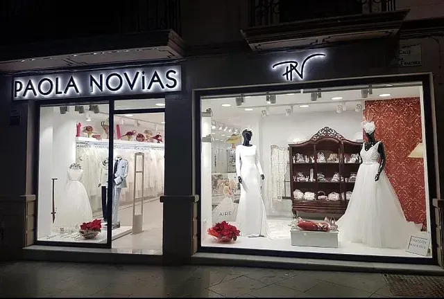 Una tienda de novias de noche, con escaparates iluminados que exhiben vestidos de novia. El letrero dice