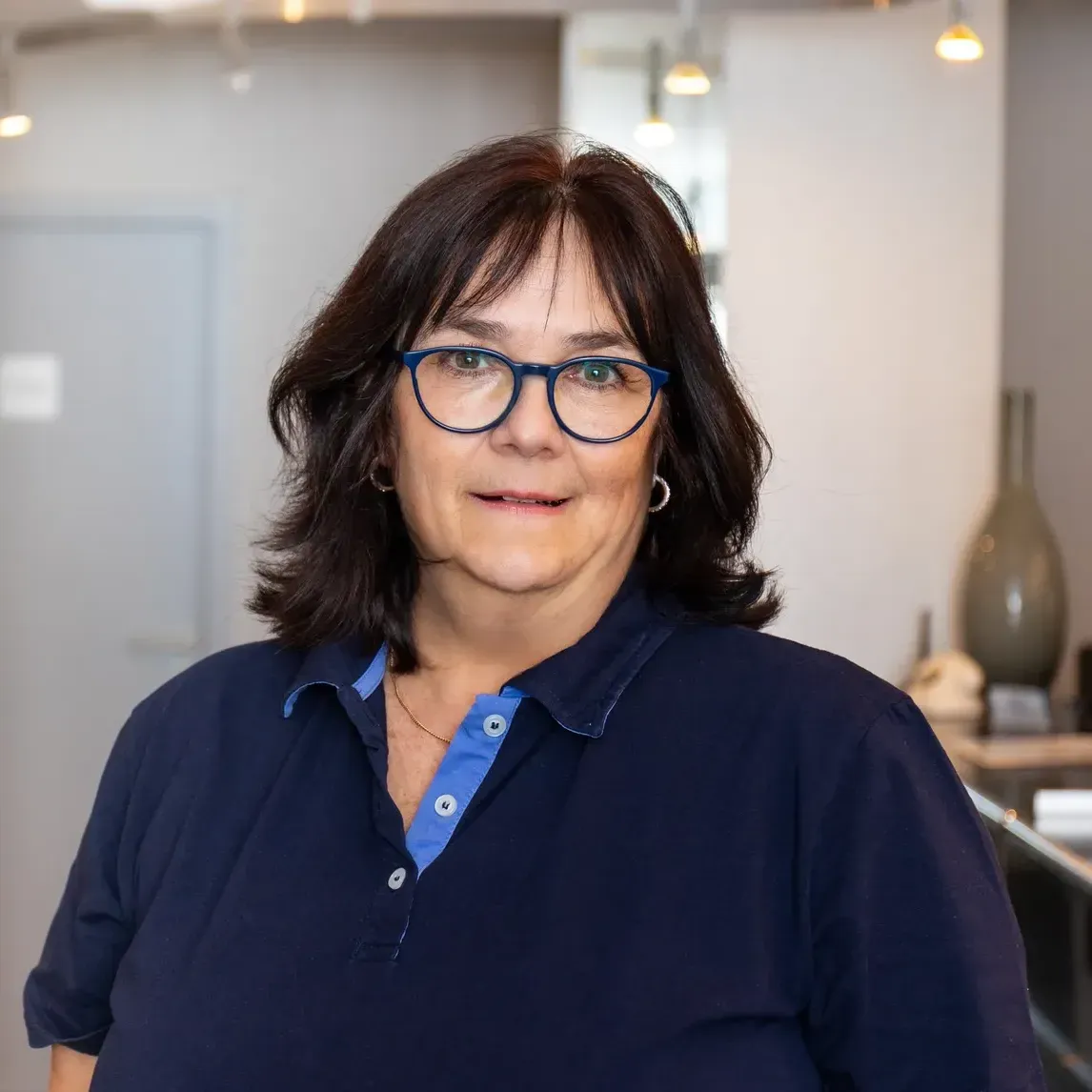 Eine Frau mit Brille und blauem Hemd steht in einem Raum.