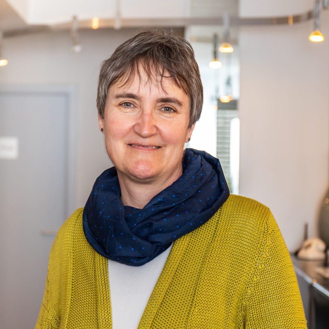 Eine Frau in einem gelben Pullover und einem blauen Schal lächelt in die Kamera.