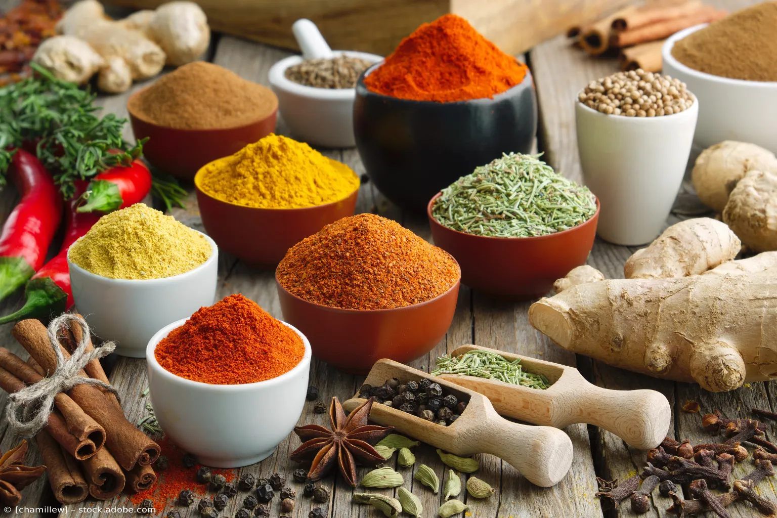 Verschiedene bunte Gewürze und Kräuter in Schalen mit Wurzeln, Samen und Chilischoten auf einem Holztisch