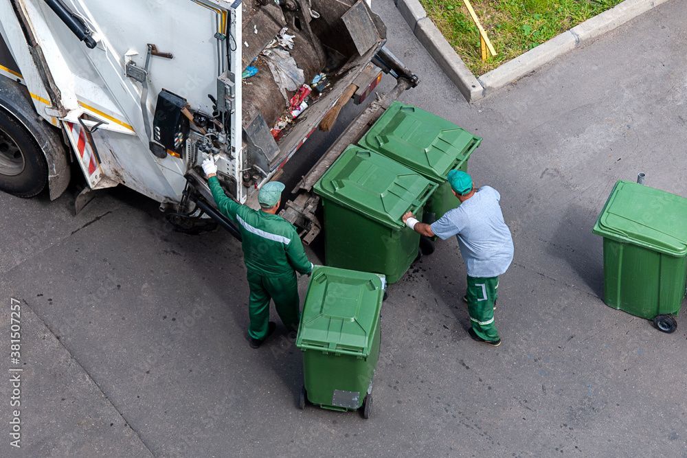 Zwei Müllwerker leeren grüne Mülltonnen in einen Müllwagen auf einer Asphaltstrasse.