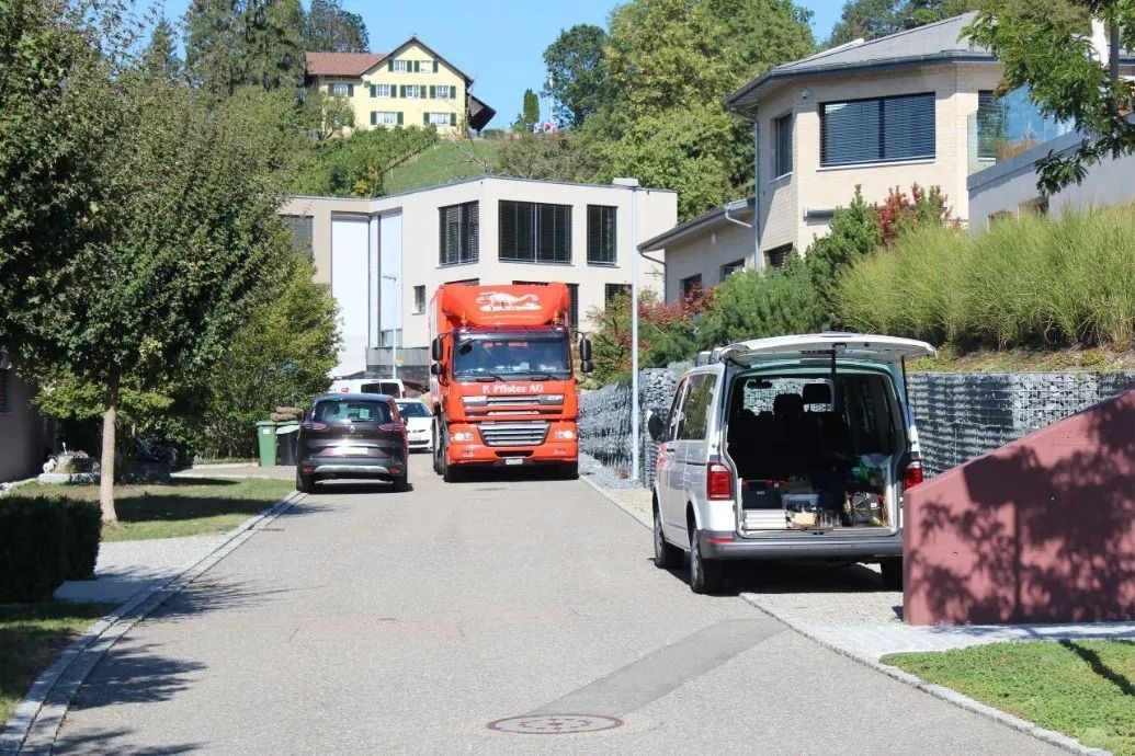 Strassenszene mit einem orangefarbenen Sattelschlepper, einem Lieferwagen und anderen Autos, die am Straßenrand geparkt sind, Häuser am Hang.