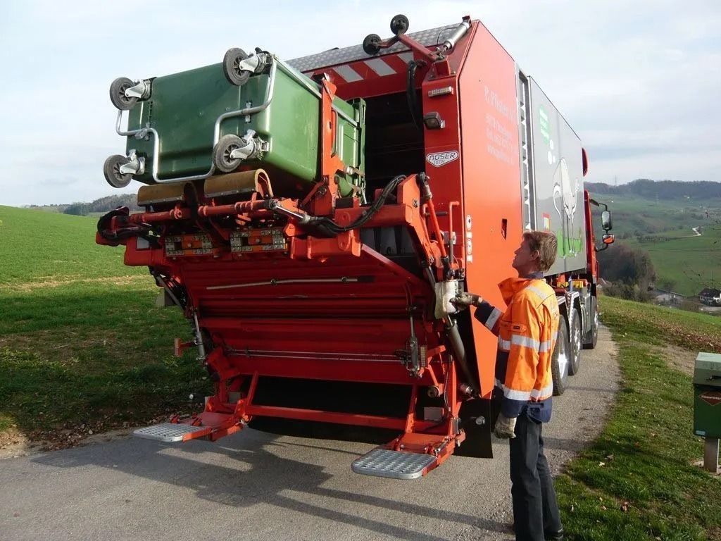 Ein Müllwagen mit einer hochgeklappten grünen Tonne; eine Person in einer orangefarbenen Weste steht in der Nähe auf einer Strasse in ländlicher Umgebung.