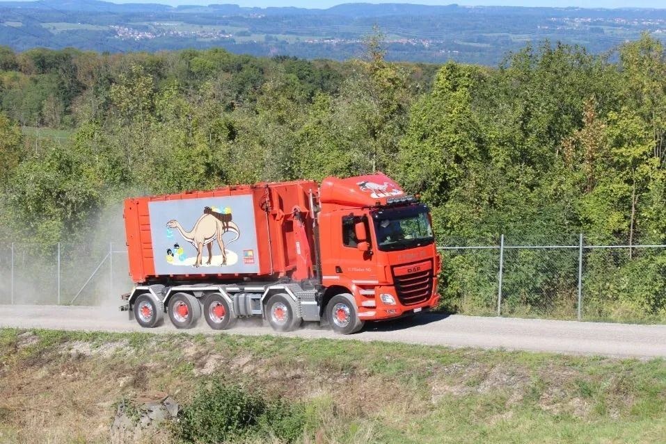 Ein roter Müllwagen fährt auf einem Feldweg; an der Seite ist eine Comicfigur zu sehen, im Hintergrund grüne Bäume.