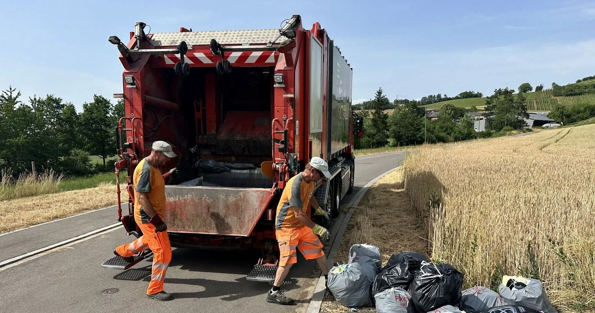 Zwei Müllwerker laden Müllsäcke in einen roten Müllwagen auf einer Landstrasse. Sonniger Tag.