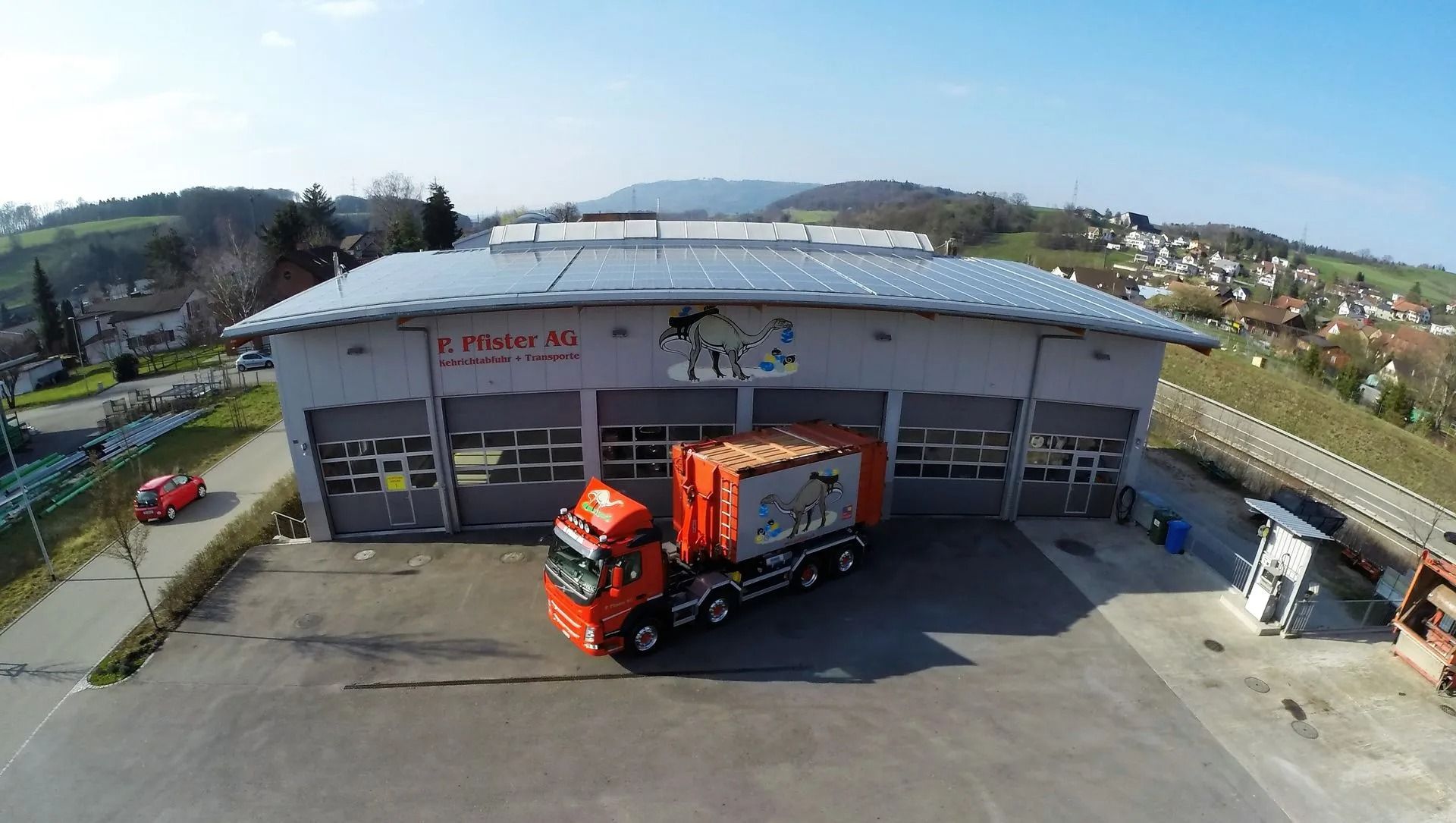 Ein orangefarbener Müllwagen vor einem grauen Gebäude mit Solardach, das sich an einen Hang schmiegt.