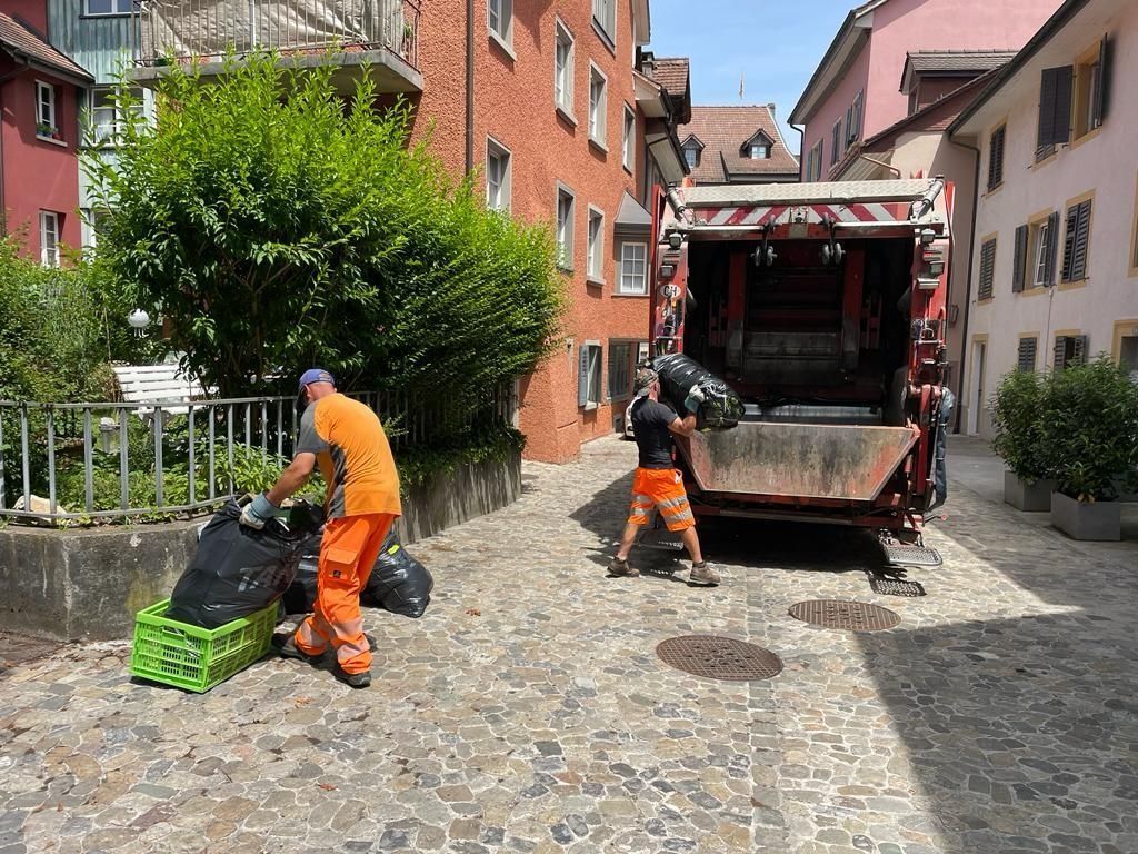 Zwei Müllwerker in orangefarbenen Anzügen laden Müllsäcke auf einer Kopfsteinpflasterstrasse in einen Müllwagen.