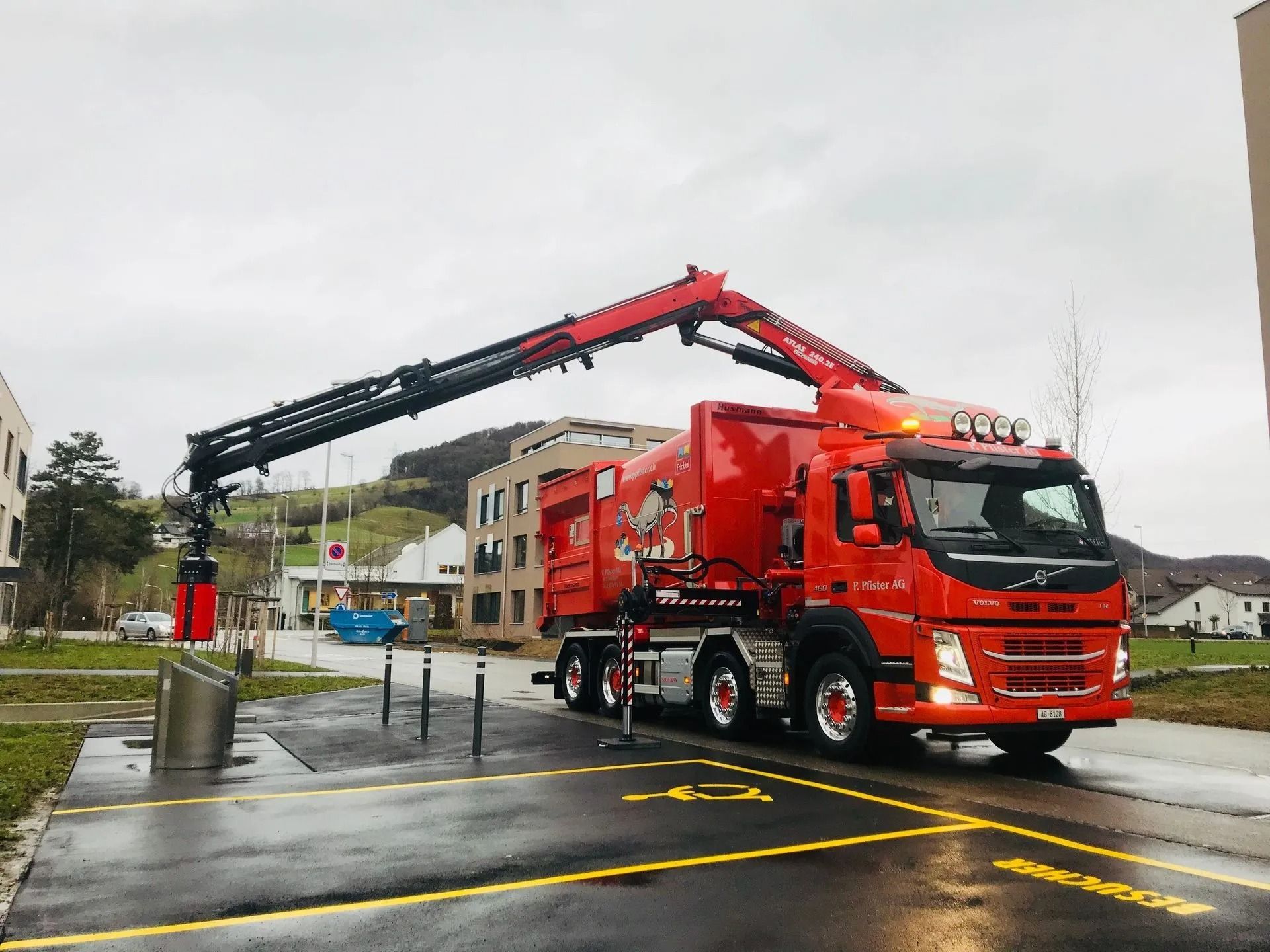 Ein roter Müllwagen mit ausgefahrenem Kranarm parkt vor Gebäuden.