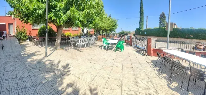 Patio al aire libre con mesas y sillas bajo los árboles, edificio rojo al fondo, día soleado.