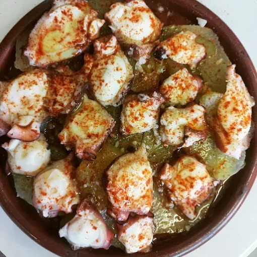 Pulpo con pimentón servido sobre patatas en rodajas, en una fuente rústica de color marrón.