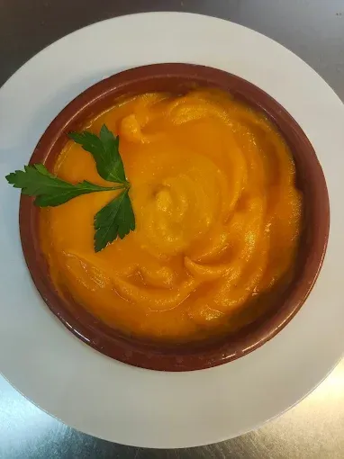 Puré cremoso de color naranja en un recipiente marrón sobre un plato blanco, adornado con una ramita de perejil.