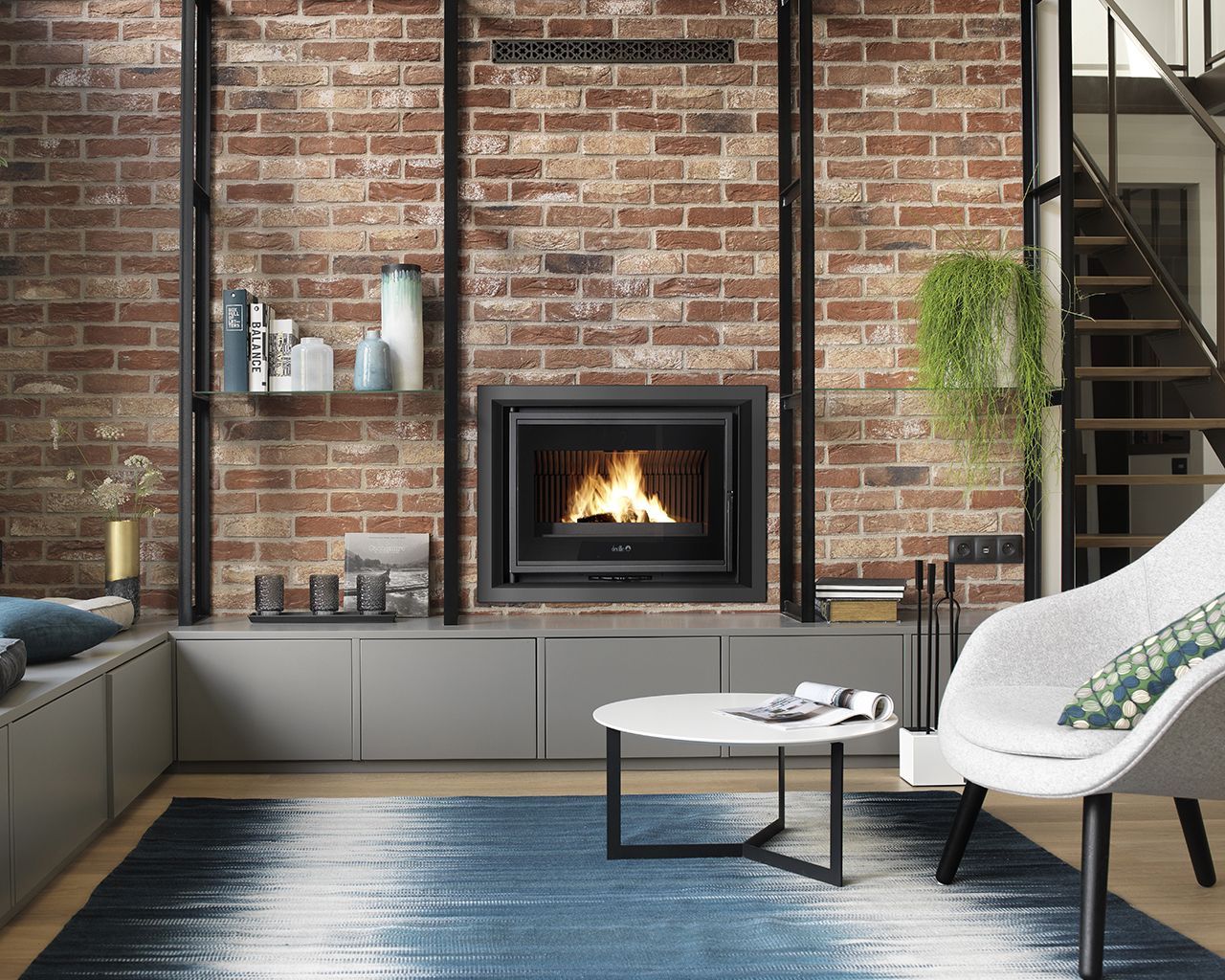 Salon moderne avec cheminée allumée, mur de briques apparentes, tapis bleu ombré, placards gris et fauteuil blanc.