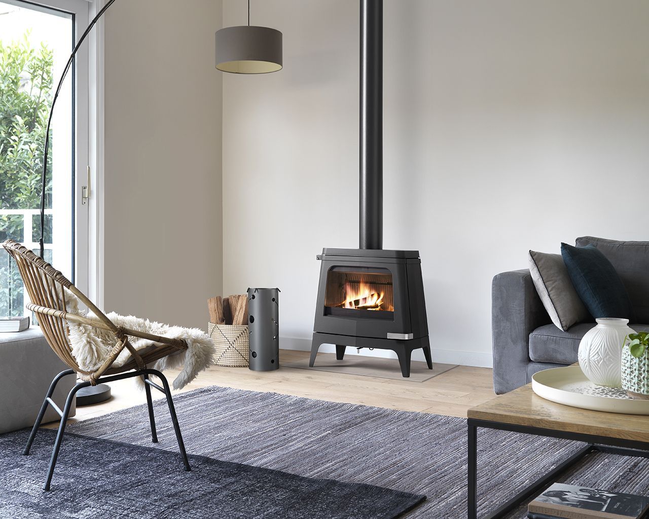 Dans un salon moderne, un poêle à bois noir avec un feu à l'intérieur est placé à côté d'un fauteuil et d'un canapé gris.
