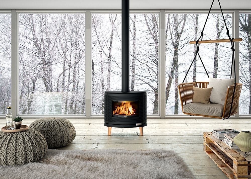 Un poêle à bois trône dans une pièce confortable aux baies vitrées donnant sur une forêt enneigée, avec un fauteuil suspendu.