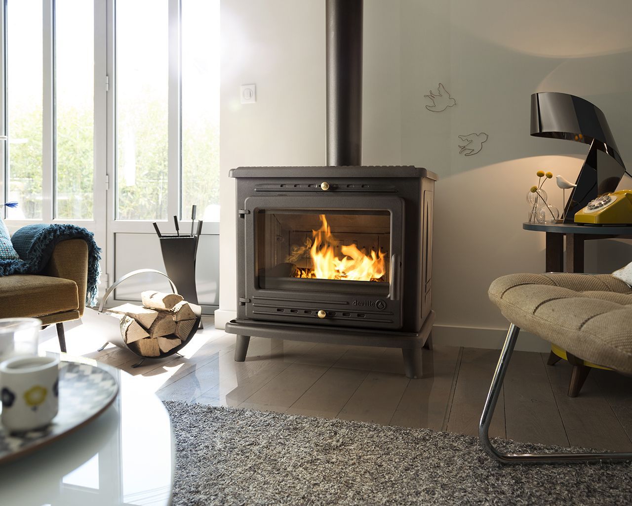 Dans un salon moderne et lumineux, un poêle à bois avec un feu allumé se dresse à côté d'un tas de bûches et de fauteuils.