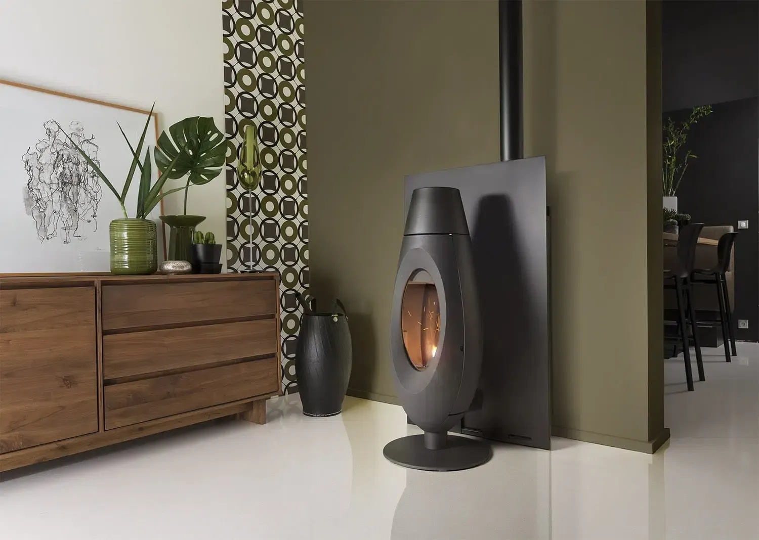 Un poêle à bois moderne aux formes sombres se dresse sur un sol blanc, devant un mur vert olive foncé orné de papier peint à motifs et de meubles.