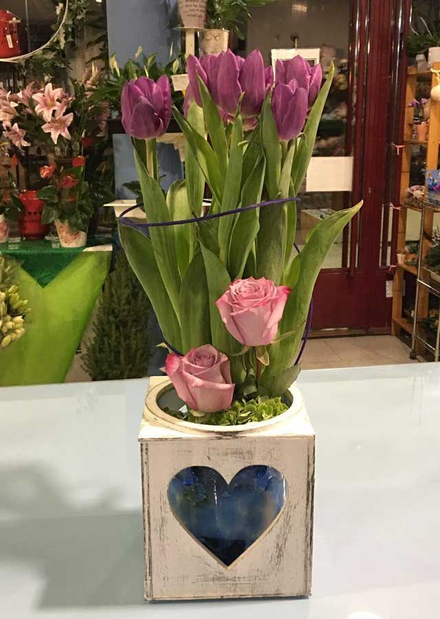 Tulipanes morados y rosas rosadas en un jarrón en forma de corazón sobre una mesa.