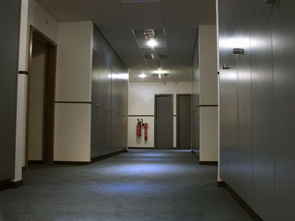 Couloir de bureau vide, murs gris, moquette bleue et extincteur rouge au fond.