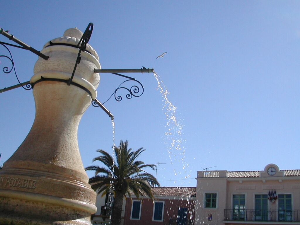 Fontaine projetant de l'eau sur une place, à côté de palmiers et de bâtiments