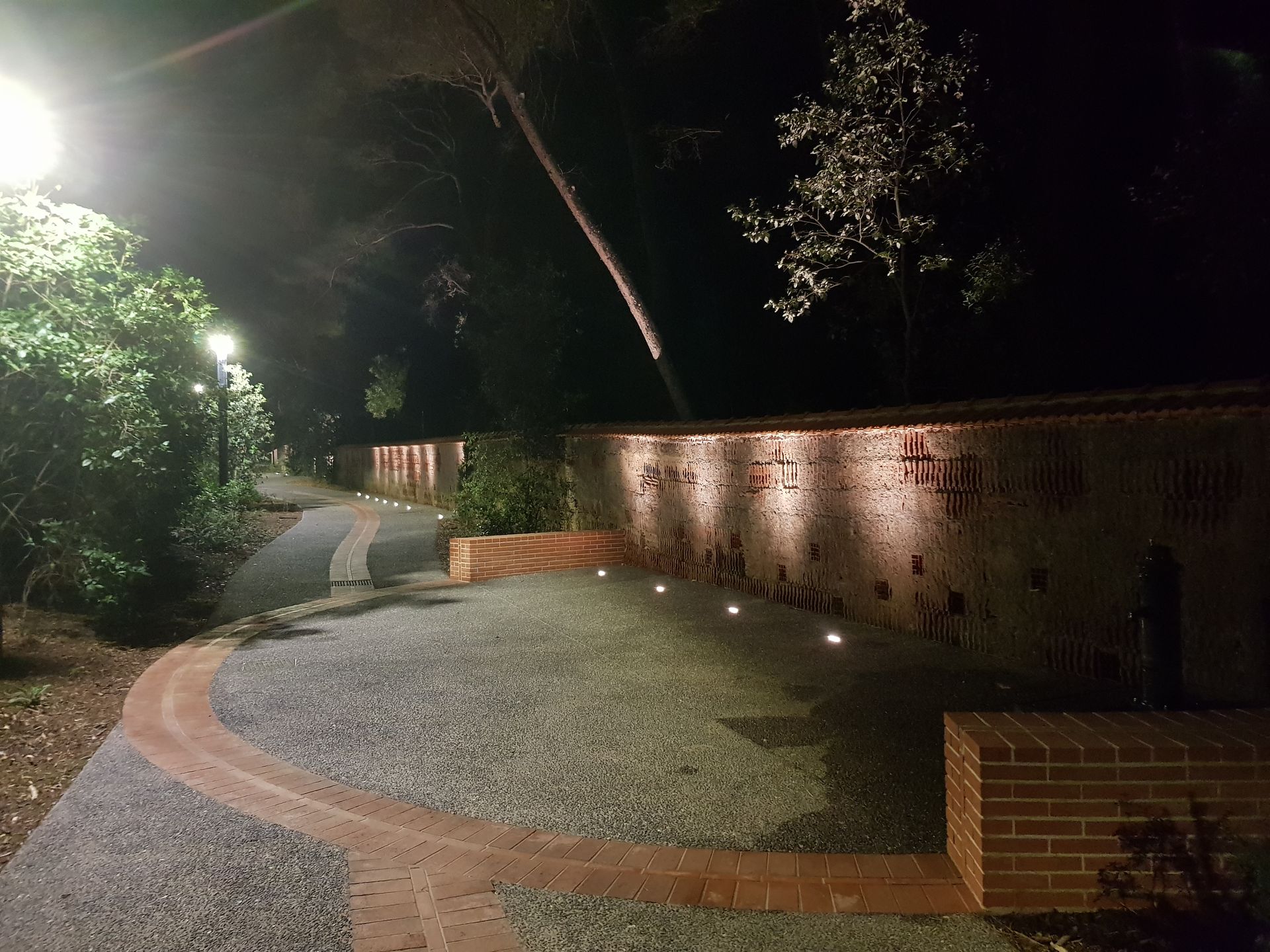 Allée nocturne de jardin avec chemin de gravier incurvé, bordure en briques, appliques murales et arbres.