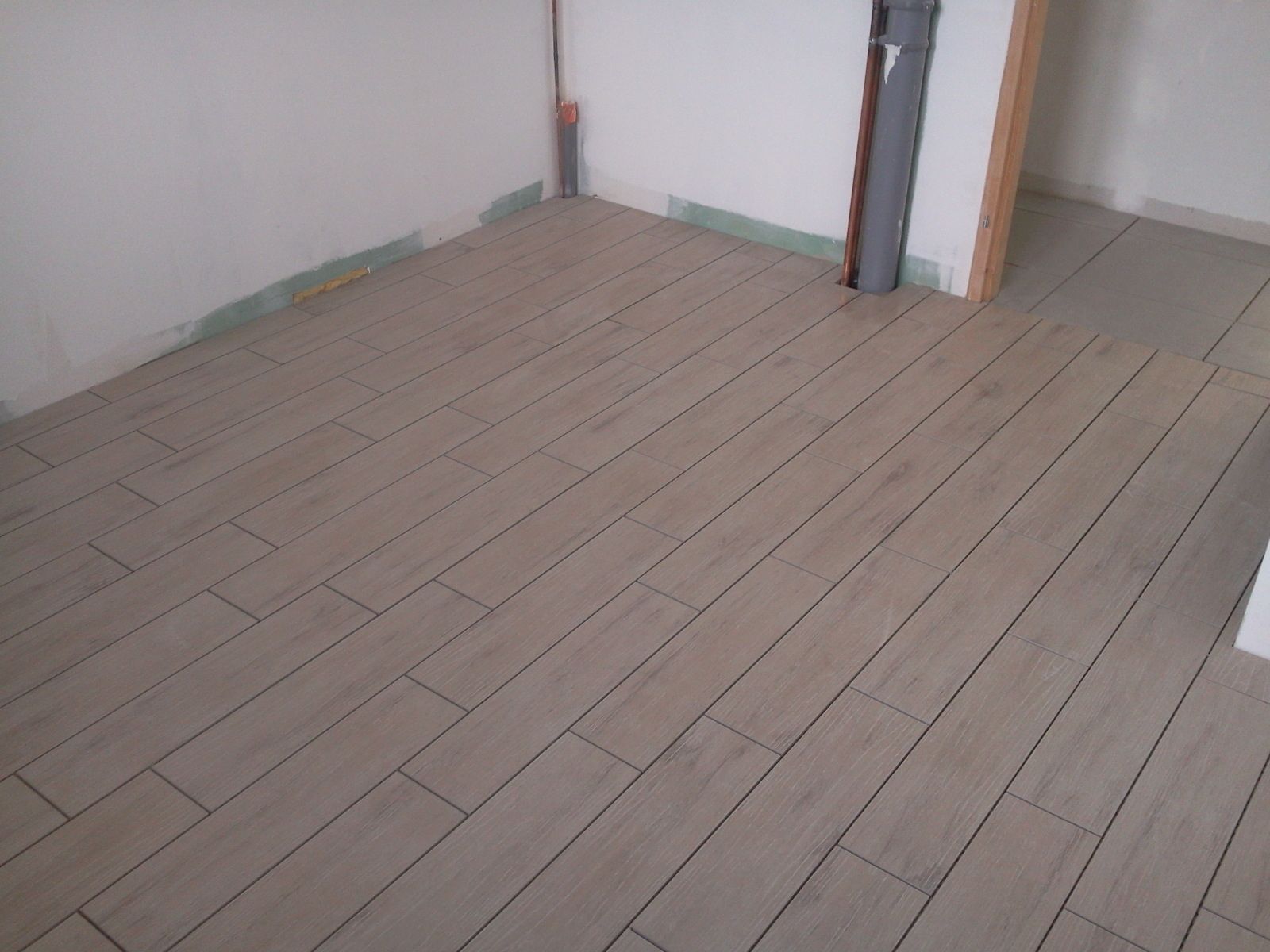 Une pièce avec des carreaux de sol rectangulaires brun clair, partiellement posés, près de murs blancs et d'une porte.