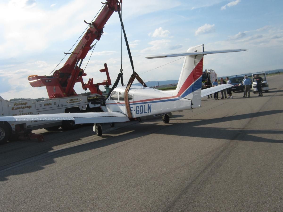 Un petit avion est soulevé par une dépanneuse rouge sur une piste goudronnée. L'avion est blanc avec des touches de rouge et de bleu.