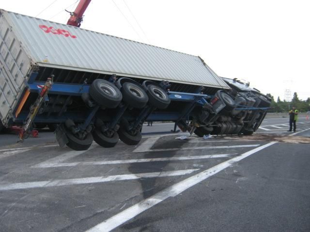 Semi-remorque renversée sur la chaussée ; conteneur de travers, pneus visibles. Une personne se tient à proximité.