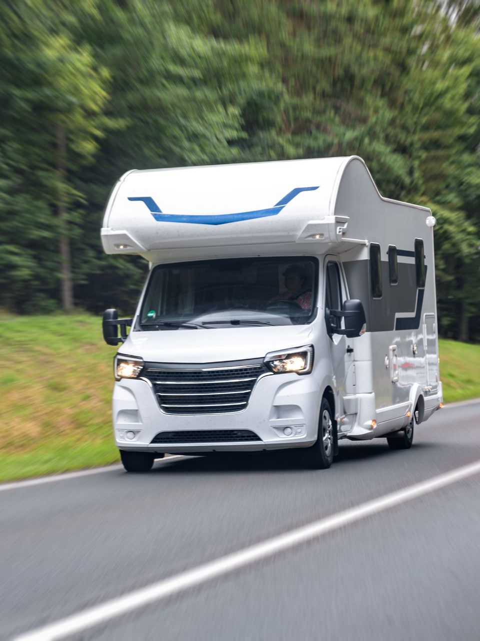 Un camping-car blanc circule sur une route goudronnée, des arbres se dressent en arrière-plan.