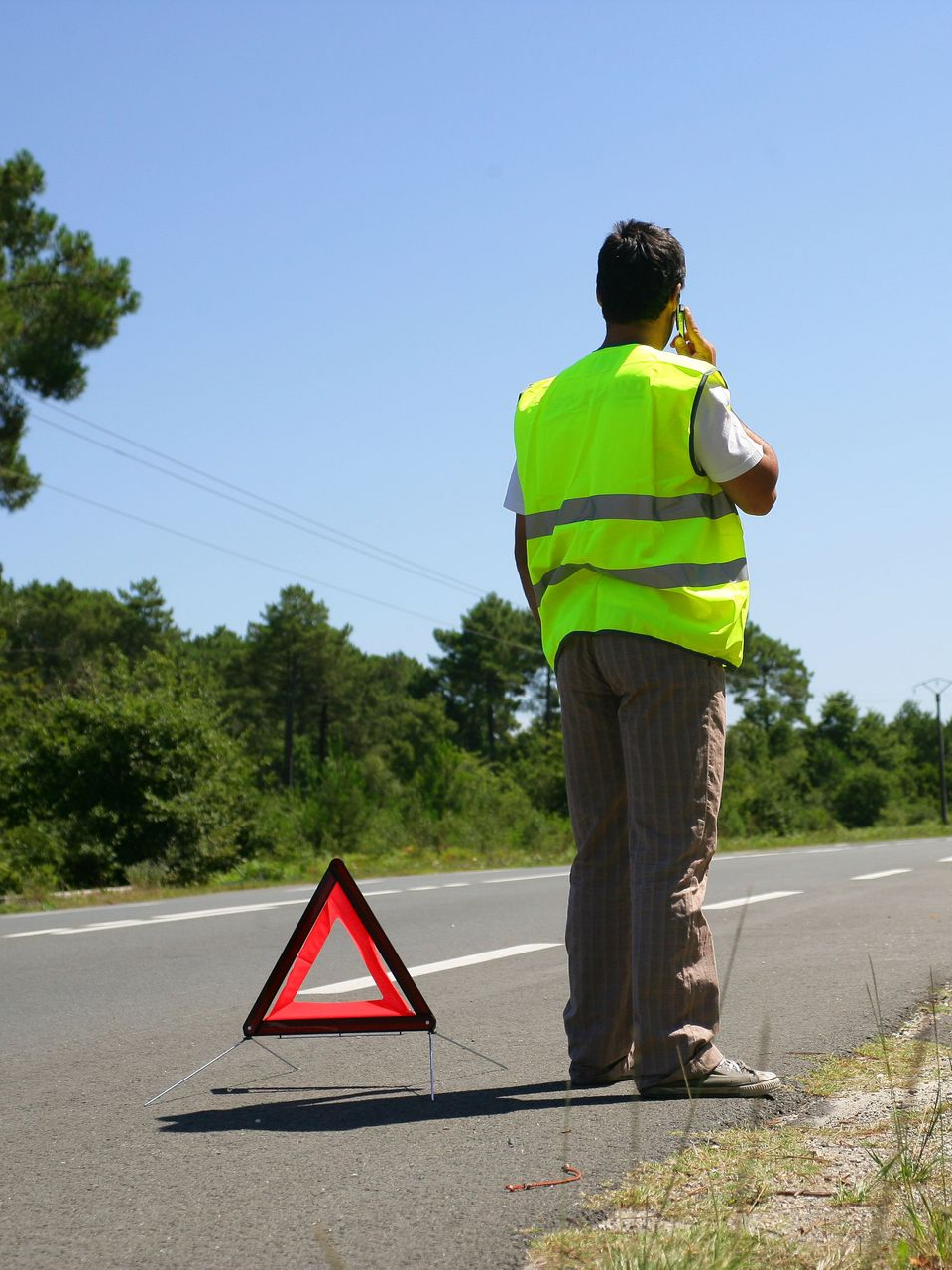 Une personne portant un gilet de sécurité jaune se trouve au bord d'une route et parle au téléphone, un triangle de signalisation étant placé au sol.