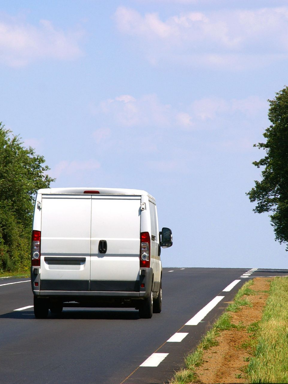 Une camionnette de livraison blanche roule sur une route noire, bordée d'arbres verts et d'herbe sous un ciel bleu.