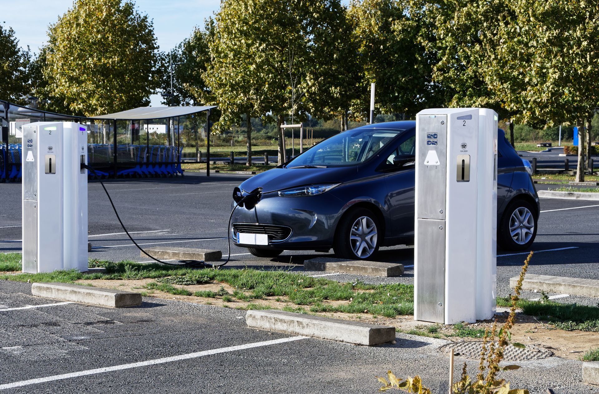 Une voiture électrique se recharge à une borne. Une voiture bleue est branchée à une borne de recharge blanche sur un parking.