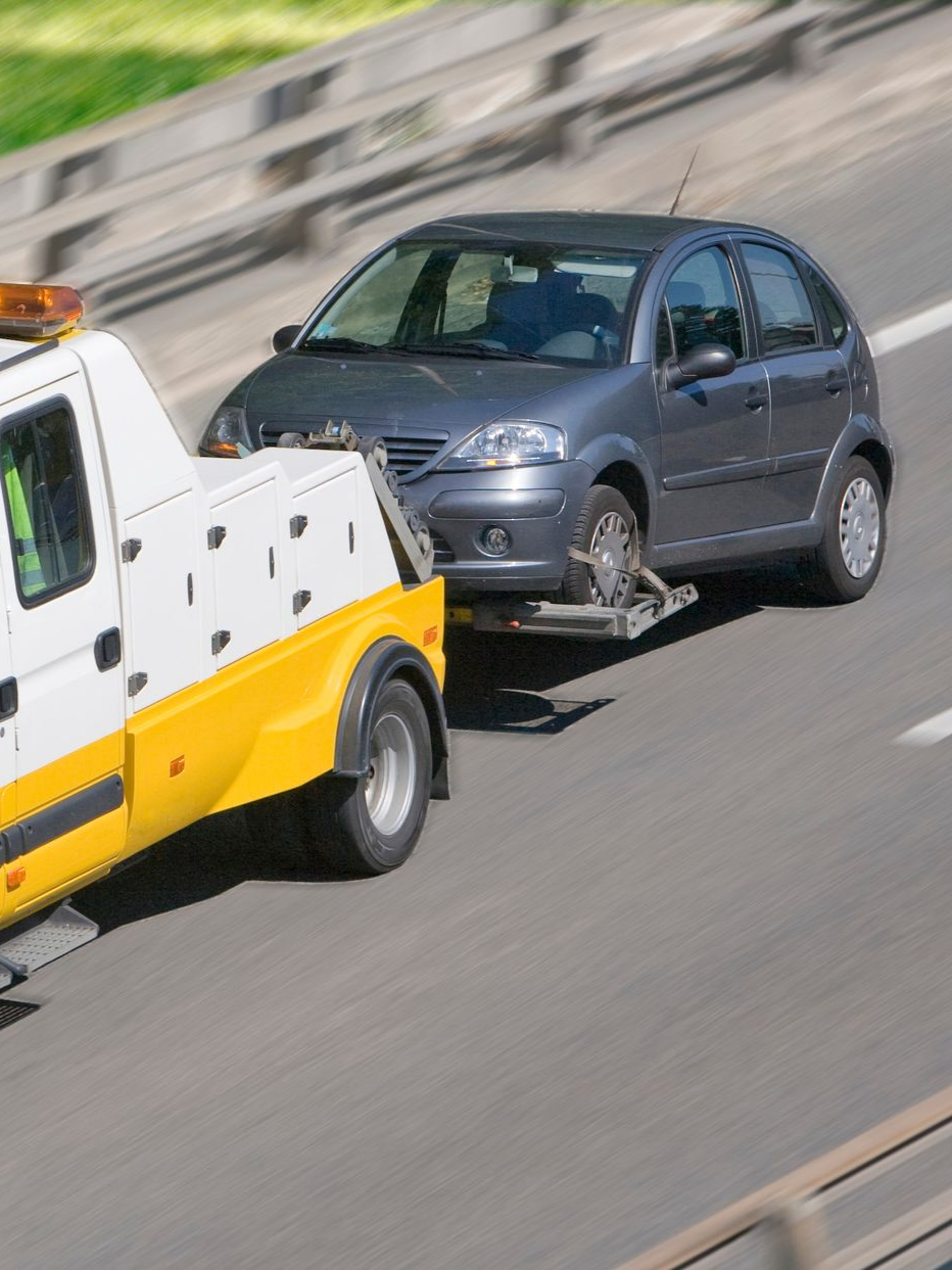 Une dépanneuse remorque une voiture grise sur une autoroute, avec un arrière-plan flou et un camion jaune et blanc.