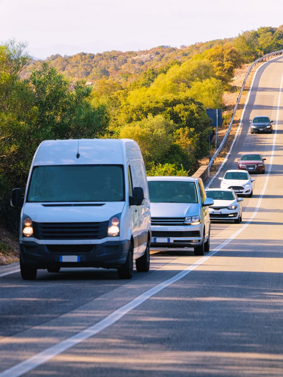 Une camionnette blanche et des voitures circulent sur une route sinueuse traversant un paysage vallonné et ensoleillé.