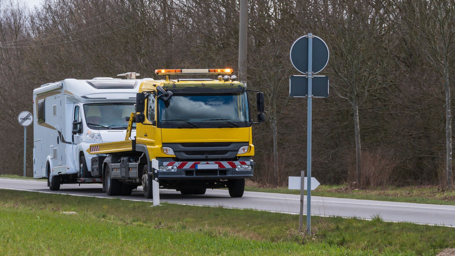 Un camion de dépannage jaune remorque une caravane blanche sur une route, feux de sécurité allumés.