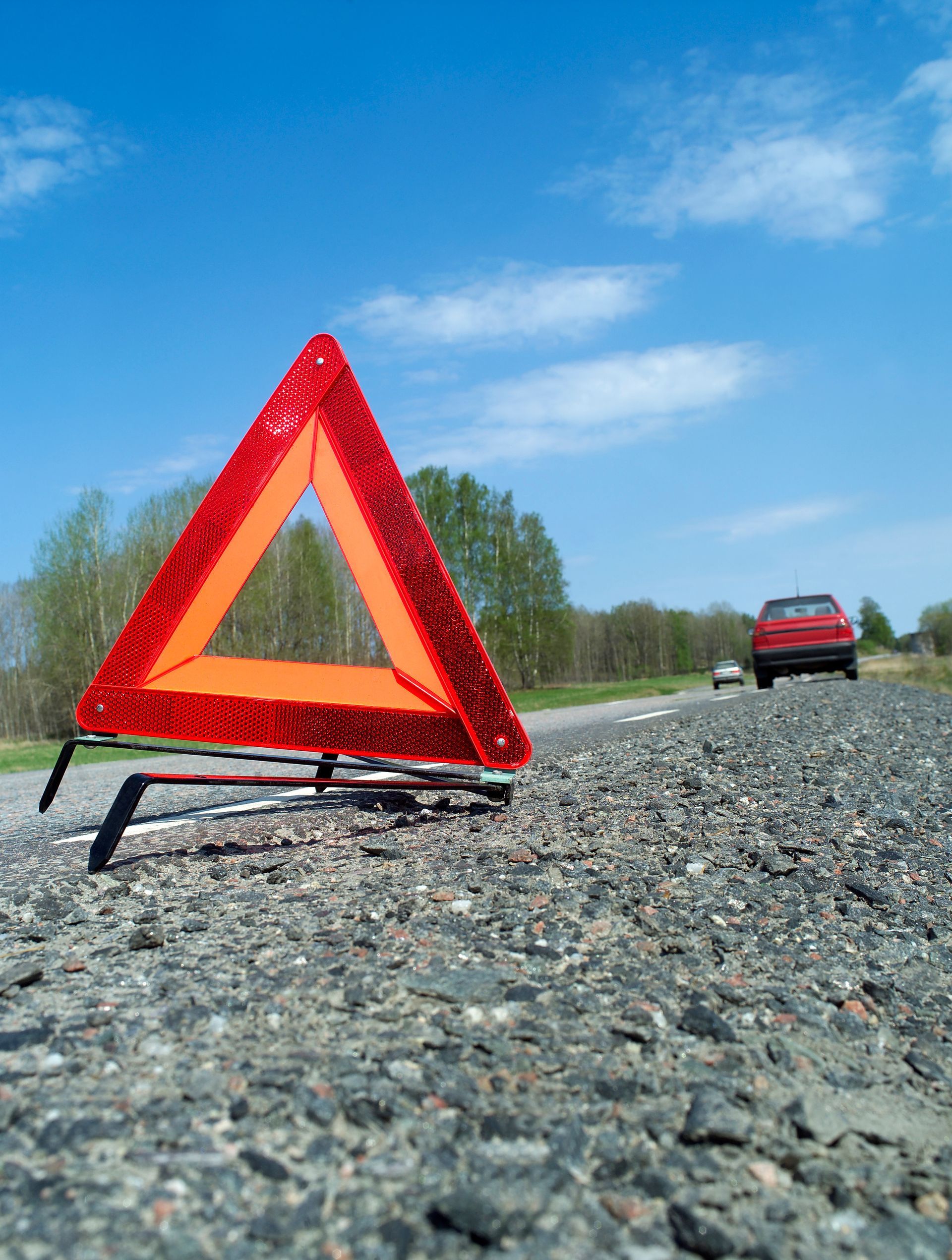 Triangle de signalisation rouge sur route asphaltée, voiture au loin, arbres et ciel bleu.