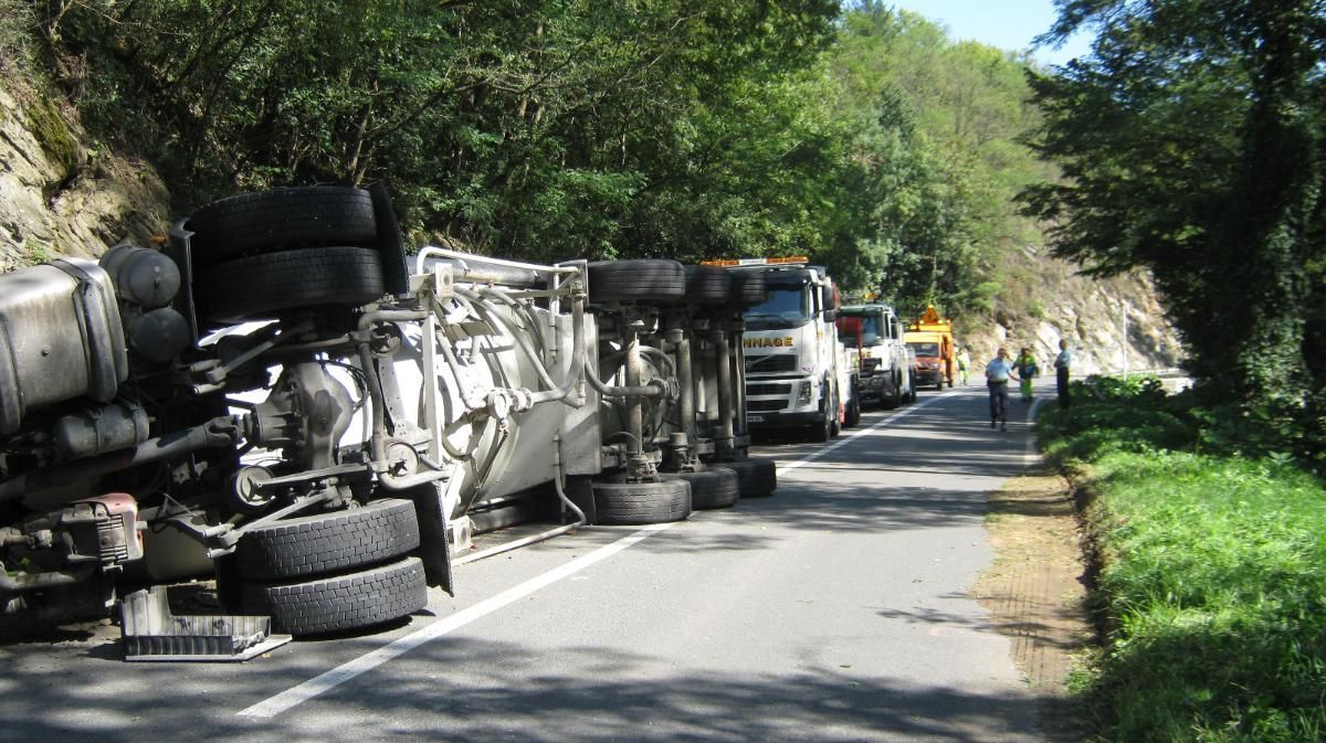 Un semi-remorque s'est renversé sur une route étroite, près d'un mur de pierre. Des véhicules de secours et des personnes sont sur place.