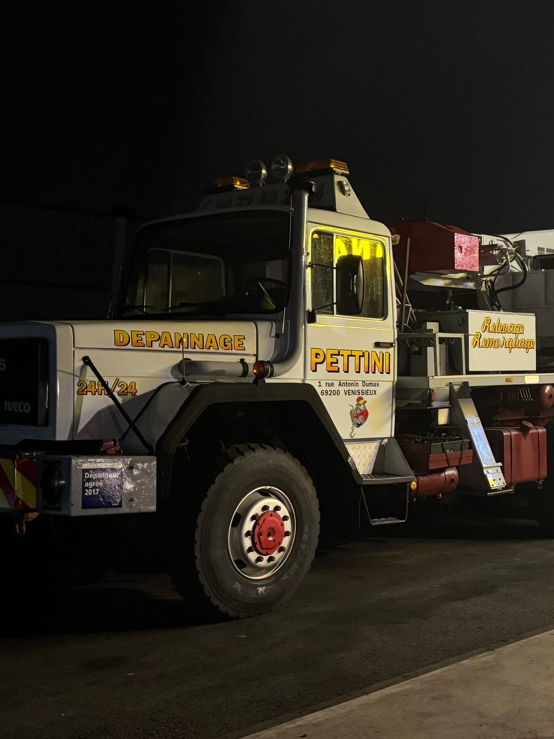 Une équipe de pose de bitume travaille de nuit sur une autoroute. On aperçoit des camions, des ouvriers équipés de projecteurs et de vêtements de sécurité réfléchissants.