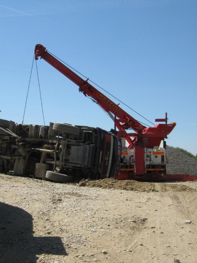 Une grue rouge soulève un semi-remorque renversé. La scène se déroule en extérieur, sur un sol poussiéreux, sous un ciel bleu.
