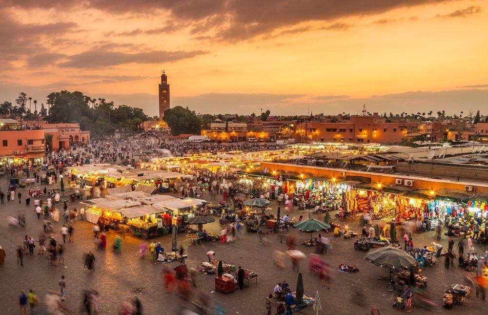 Sunset view of Djemaa el-Fna square, Marrakech, Morocco, bustling with people, vendors, and lit stalls.