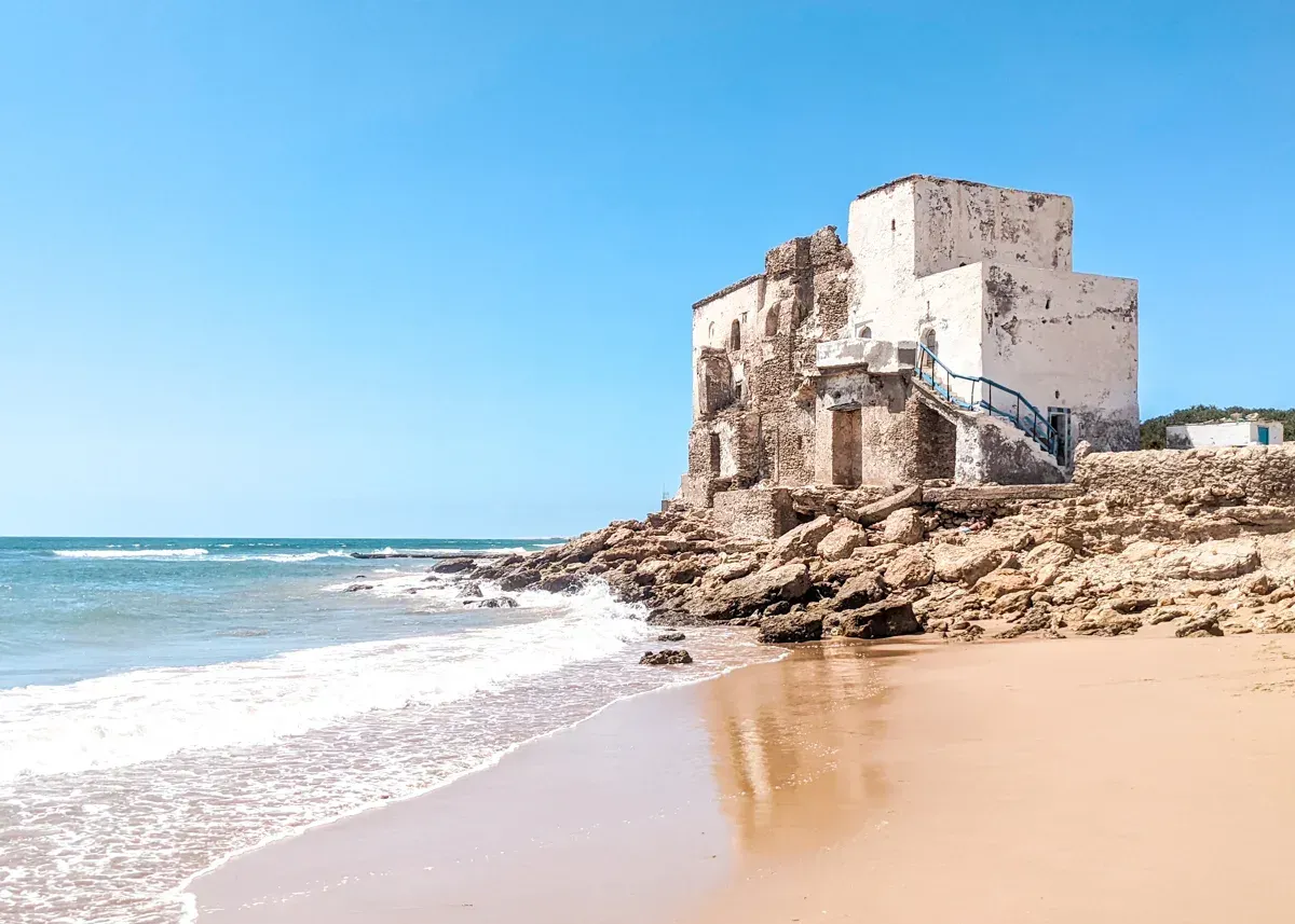 Old white building in Sidi Kaouki on a rocky shore, waves crashing onto the sandy beach under a blue sky.
