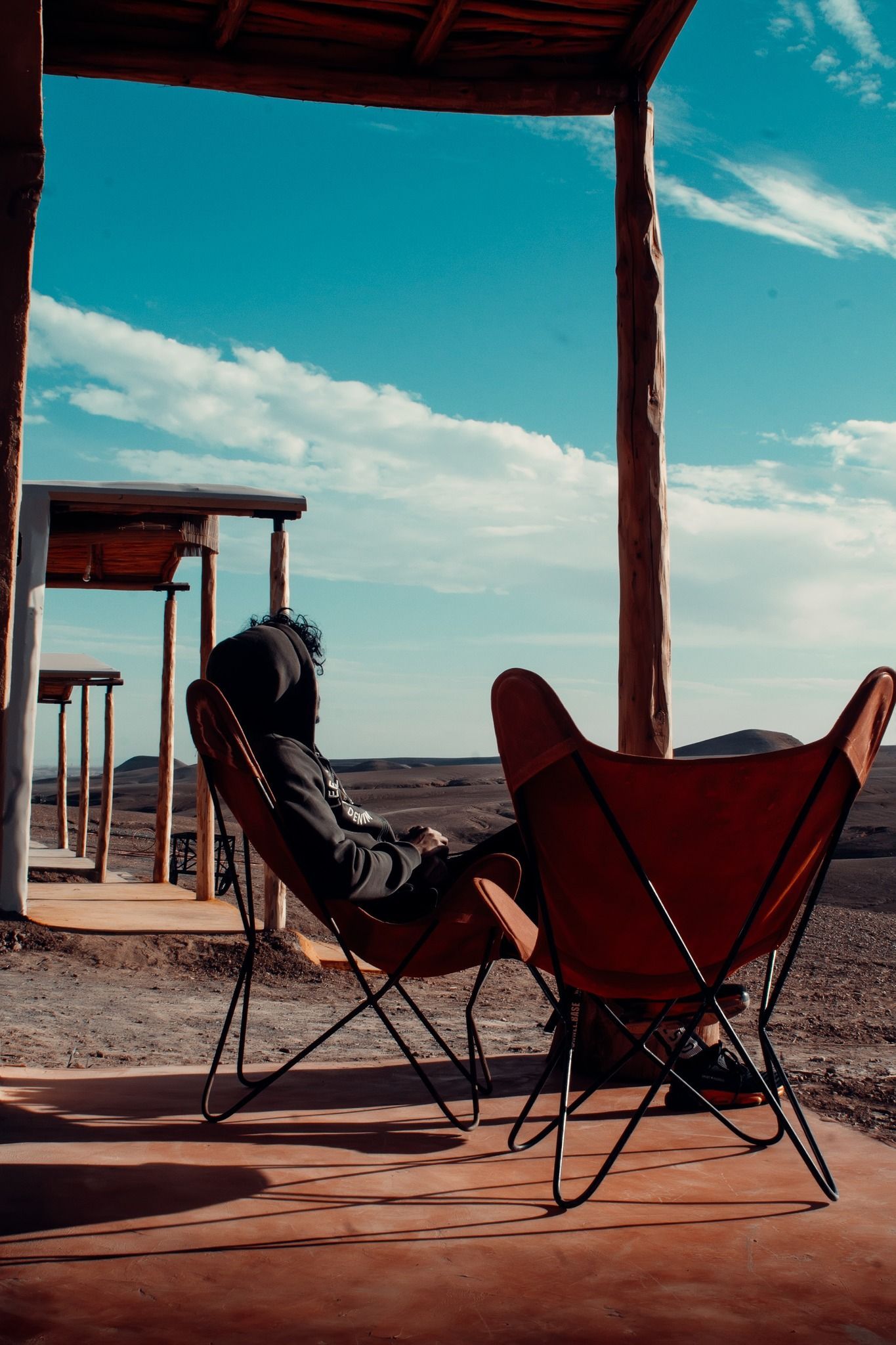 Person relaxing in a butterfly chair on a porch, overlooking a desert landscape under a bright blue sky.