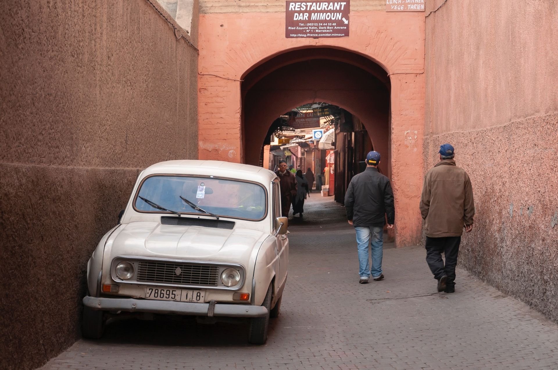 A white car parked under an arched doorway in a narrow street in Morocco; two men walk ahead.