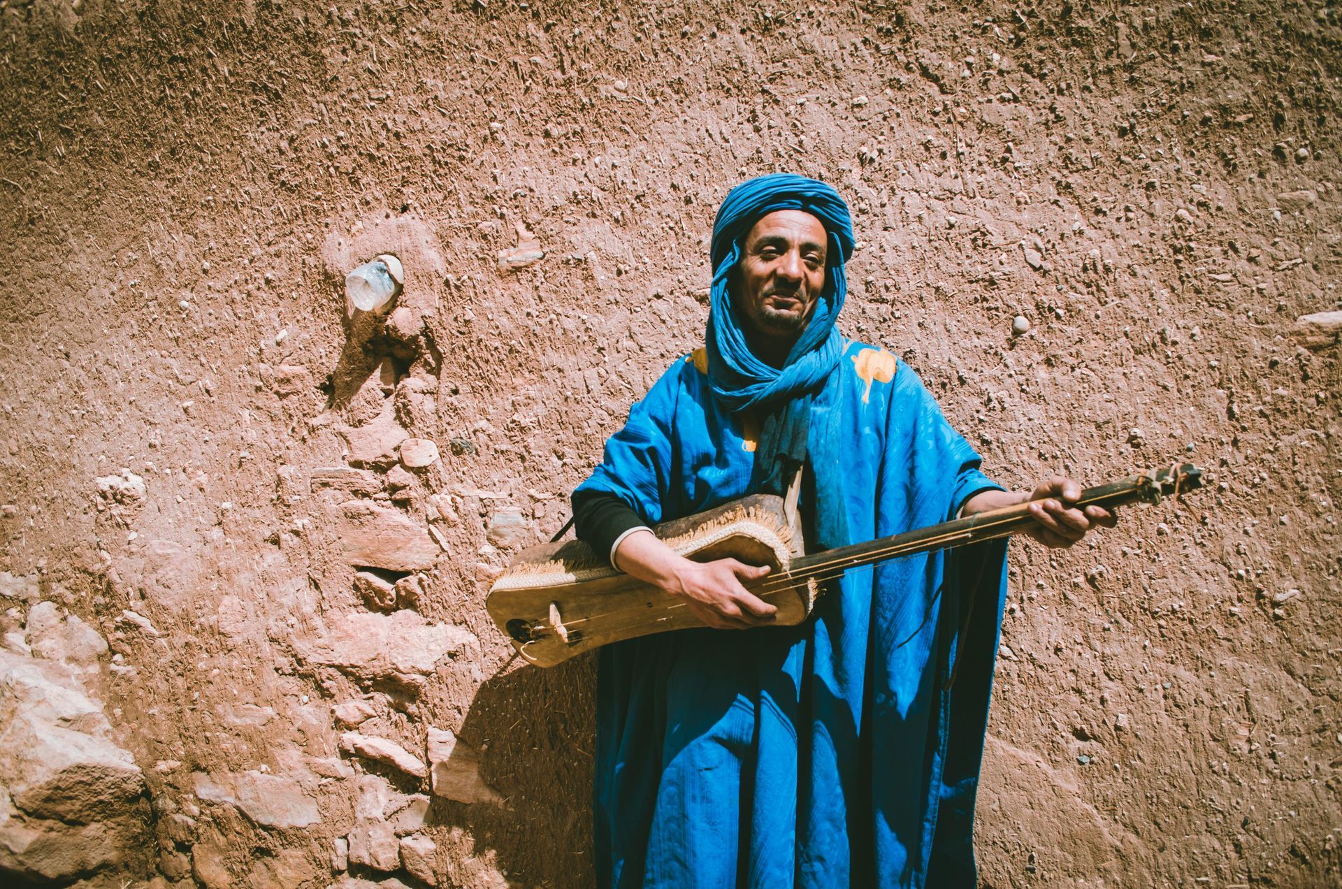 Man in blue traditional clothing playing a stringed instrument against a rustic, earthy-colored wall.