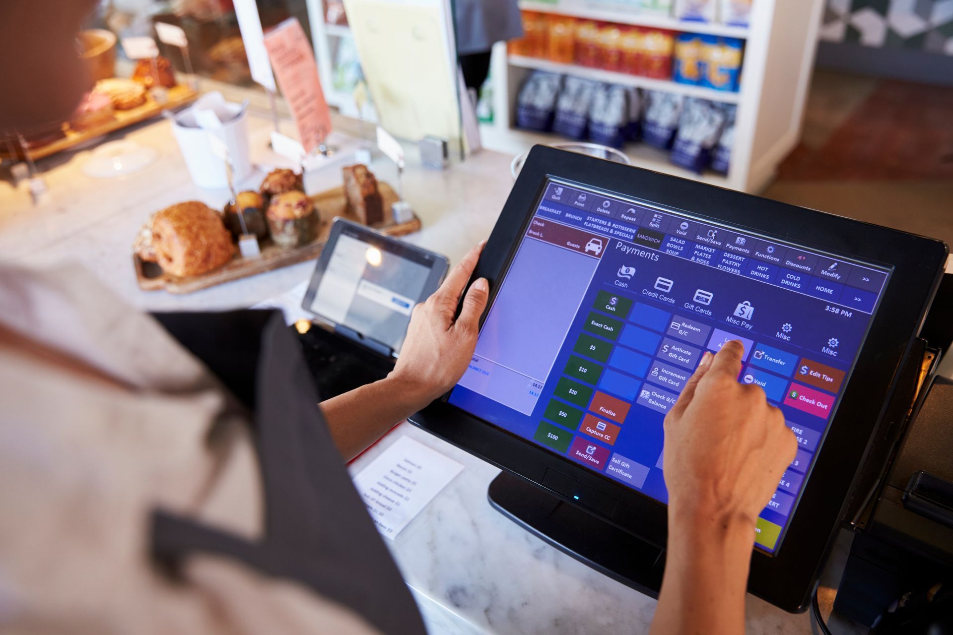 Une personne utilise une tablette tactile de point de vente au comptoir d'un café, avec des pâtisseries et une carte de crédit à proximité.
