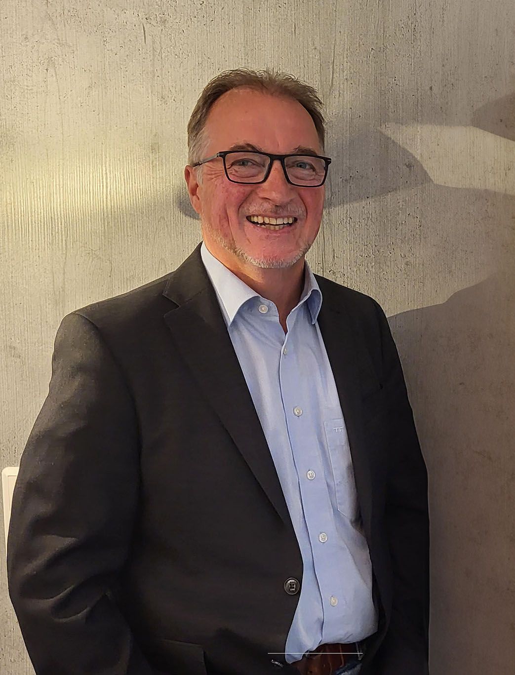 Ein Mann im Anzug und mit Brille steht lächelt vor einer Wand.