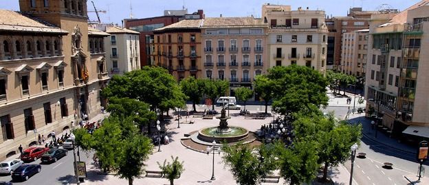 Una plaza en Valencia, España, con árboles, una fuente y edificios a su alrededor. Se ven algunos vehículos y peatones.