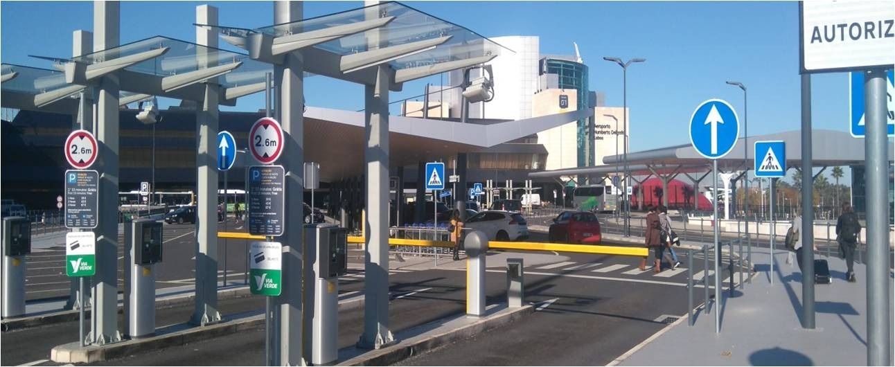 Una entrada al aeropuerto con cabinas de peaje, señalización aérea y señales de tráfico que orientan a los conductores. Cielo azul despejado al fondo.