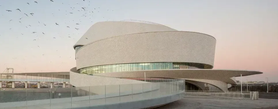 Un edificio moderno, blanco y en espiral, con ventanas de cristal. Una pasarela curva conduce a la entrada. Pájaros vuelan en el cielo.