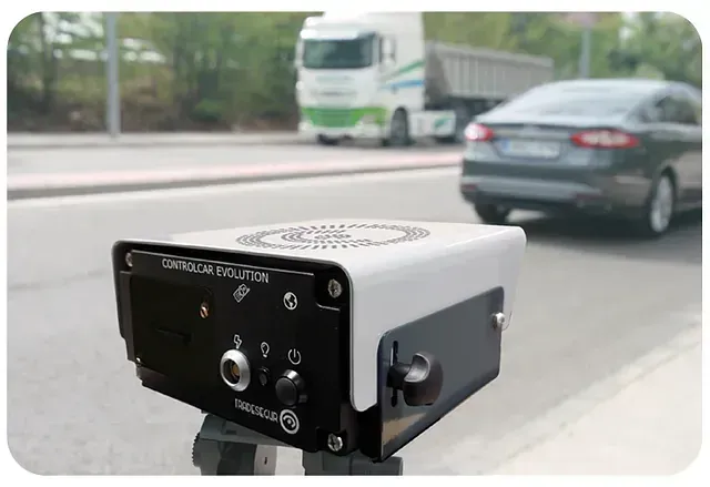 Dispositivo de control de velocidad sobre un trípode, enfocado en vehículos y un camión que pasan por la carretera. El dispositivo es blanco y negro.