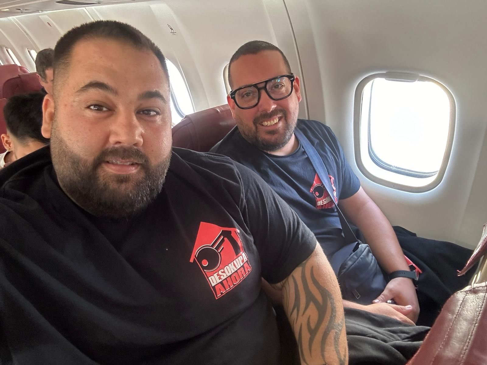 Dos hombres están sentados en un avión y uno de ellos tiene un tatuaje en el brazo.